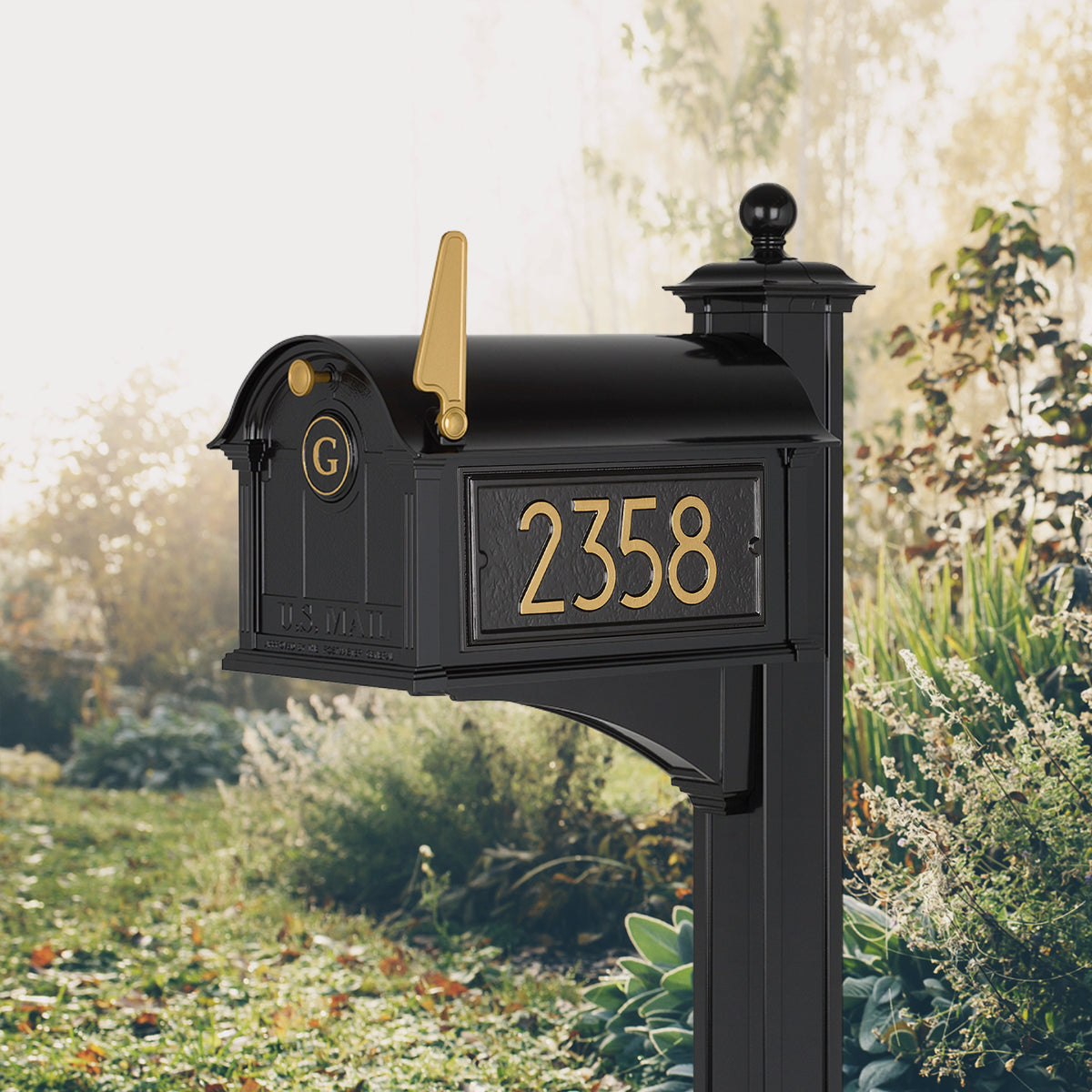 Modern Balmoral Monogram Mailbox & Post Package in black and gold, shown beside a lawn with sample address numbers and monogram
