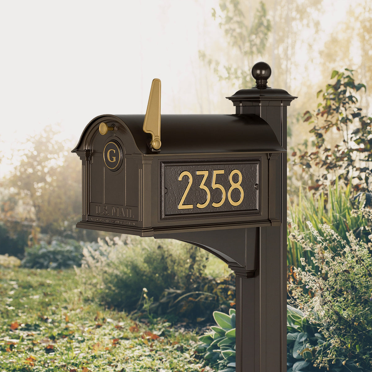 Modern Balmoral Monogram Mailbox & Post Package in dark bronze and gold, shown beside a lawn with sample address numbers and monogram